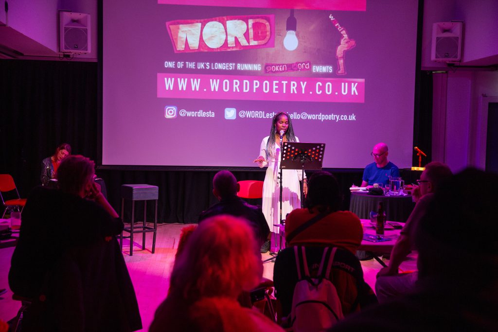 A woman in front of a purple projected screen for a WORD! event, speaking to an audience.