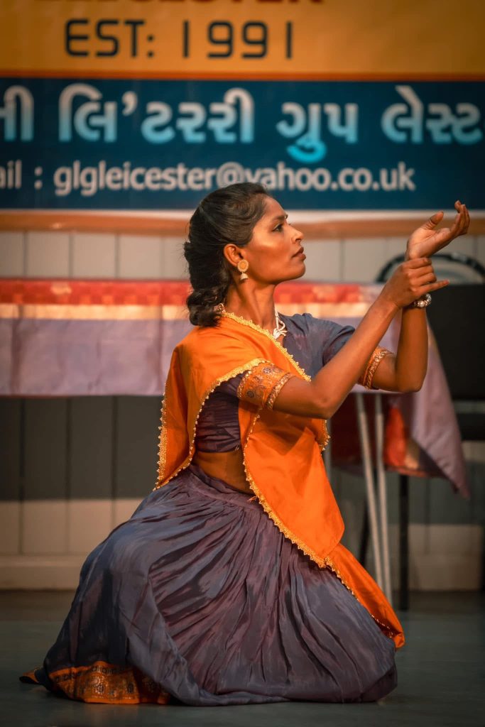 An Asian women in traditional Indian dance clothes, dancing in front of a striped background.