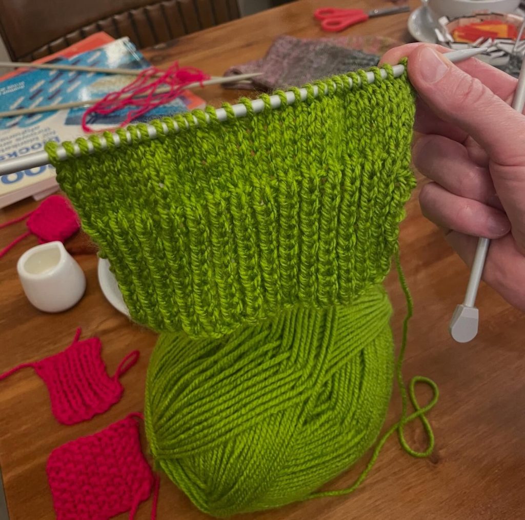 A hand holding up a knitting needles with rows of green stitches falling down towards a ball of yarn.