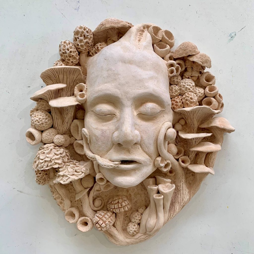 A clay sculpture of a face surrounded by mushrooms and fungus.