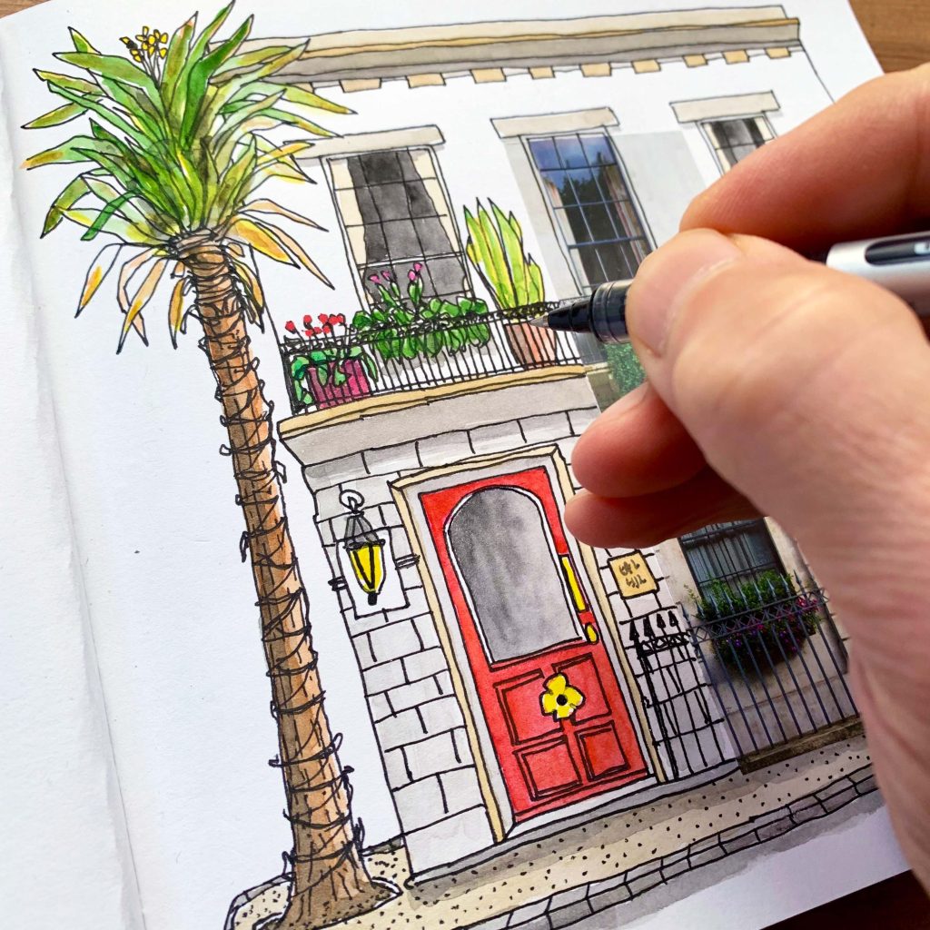A hand holding a pen drawing a colourful English house with a tree outside.