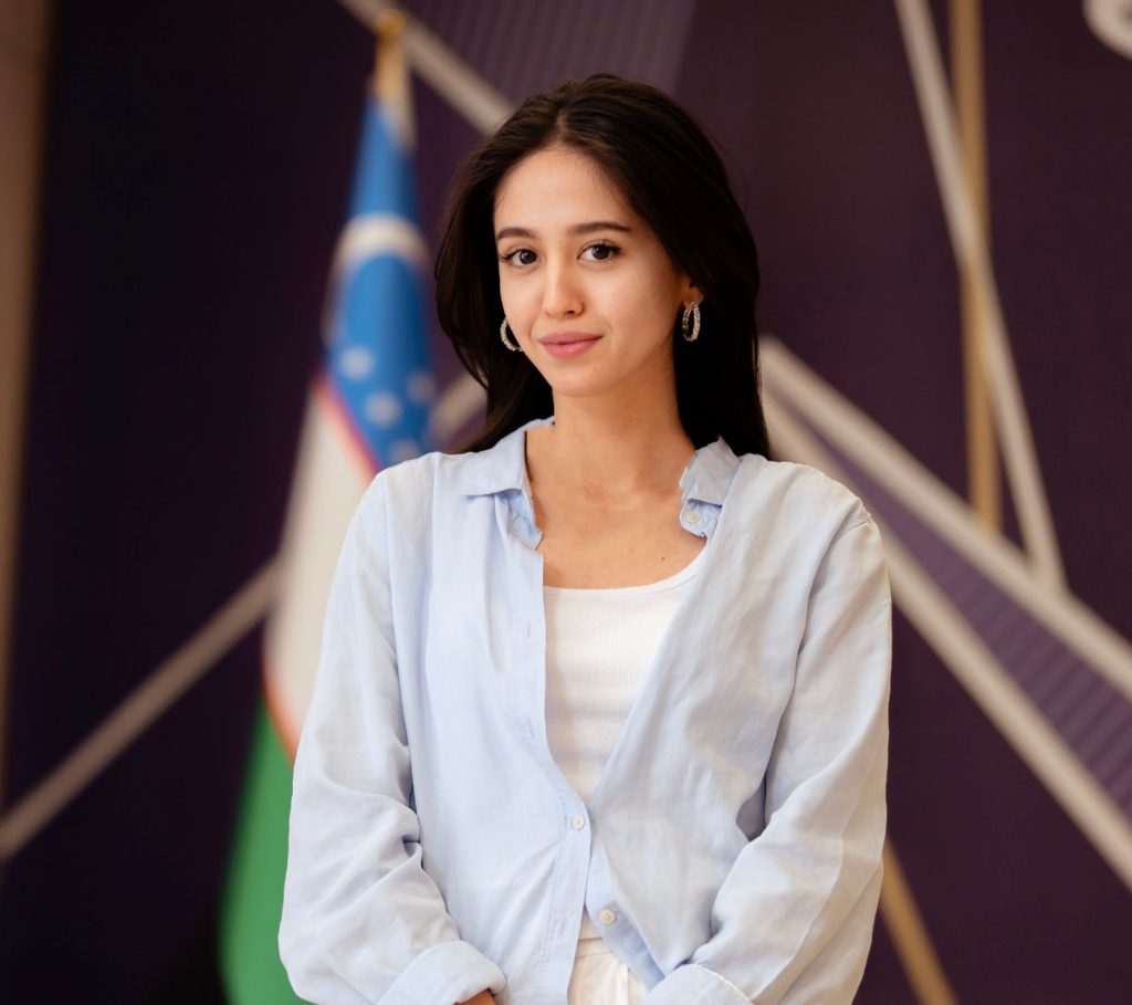 A young woman with dark hair wearing a white vest top and light blue button-up shirt, standing in front of a blurry flag and abstract wall.