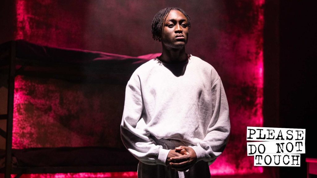 A Black man in a grey sweatshirt standing in front of a pink and black textures background. In the bottom right corner is says 'Please do not touch'.