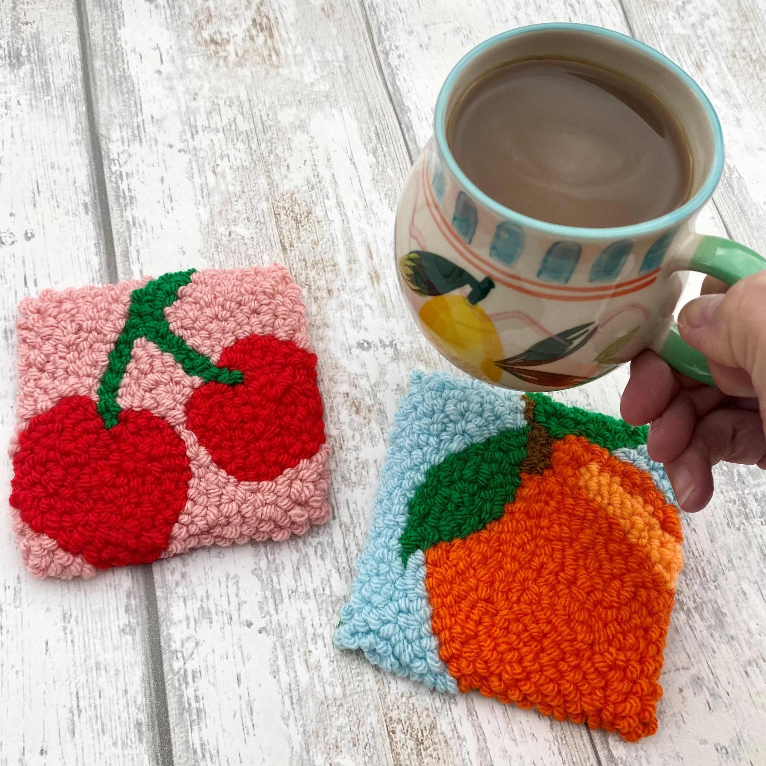 Two punch needle coasters of a cherry and a orange, with a mug about to be placed onto it.