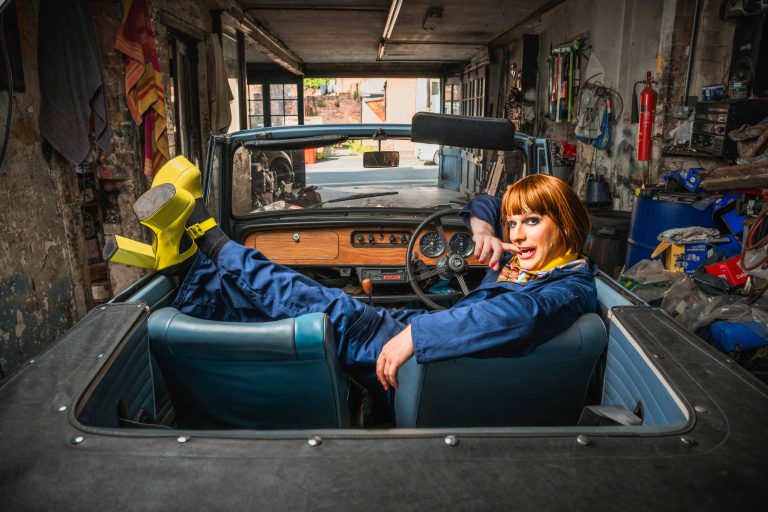 A red headed person in a blue boiler suit and yellow heels, laying in a open-top car in a mechanics station.
