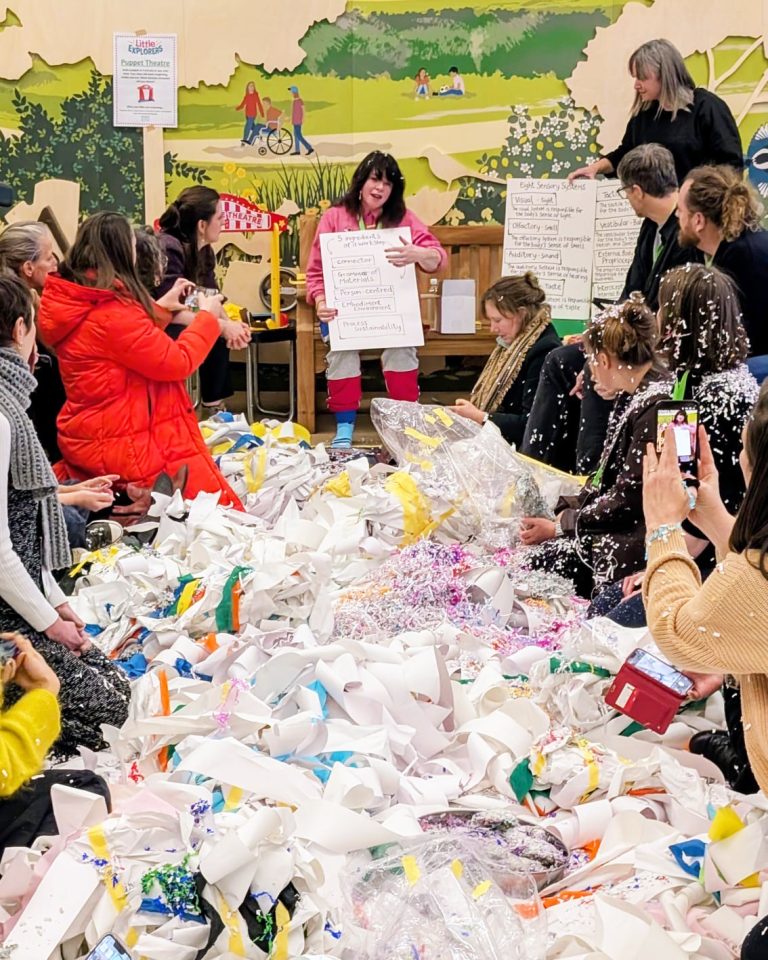 A room full of cut up paper and creative materials in a heap on the floor, surrounded by people listening to a woman at the top holding a giant piece of paper.