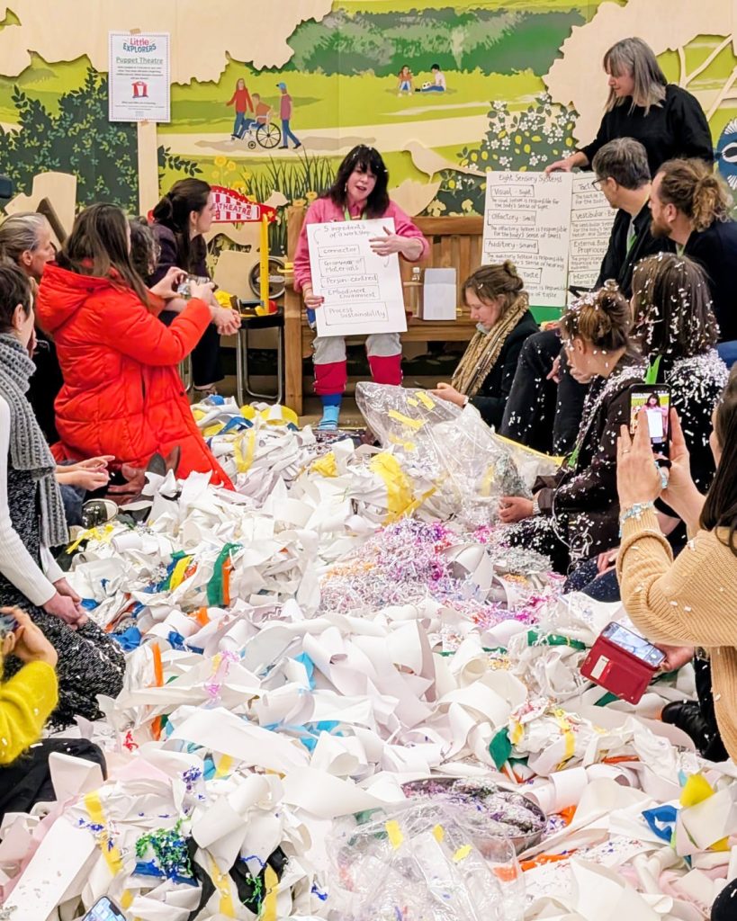A room full of cut up paper and creative materials in a heap on the floor, surrounded by people listening to a woman at the top holding a giant piece of paper.