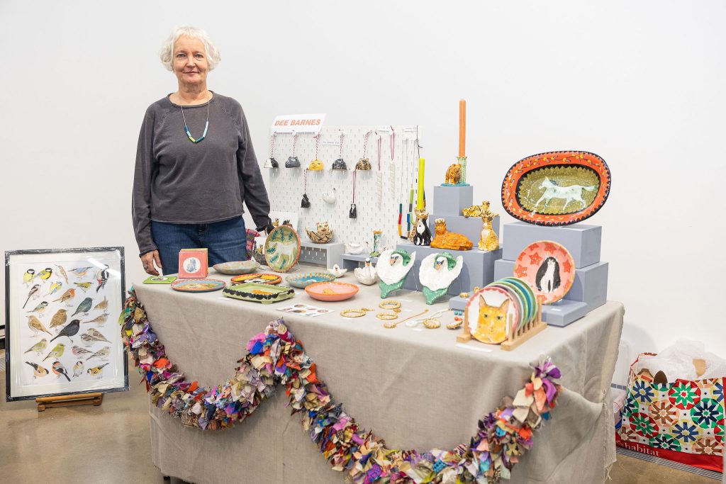 An older women with white hair in a grey jumper stood next to a table filled with colourful ceramics she is selling.