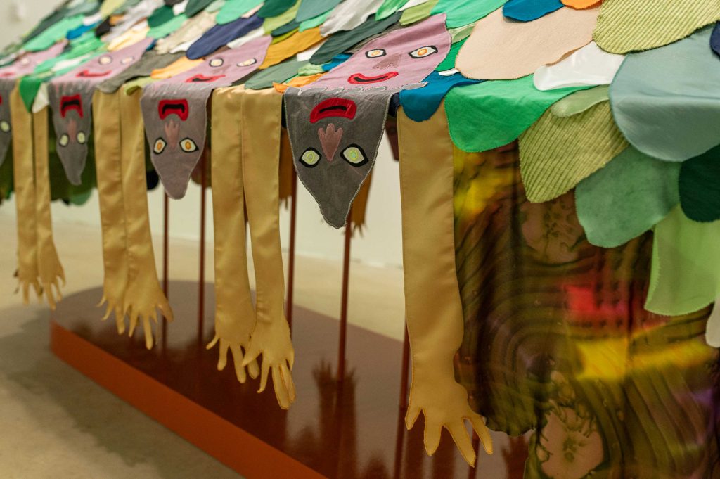 A sculpture made of fabrics showing five pink heads on top of a scaled mound on a table. From the table drips down the faces and hands of more people. From each side is a large green flower with a shocked face going upwards.