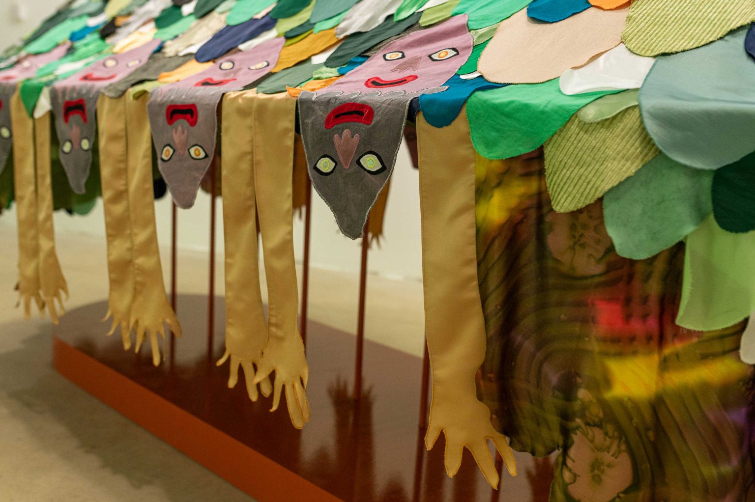 A sculpture made of fabrics showing five pink heads on top of a scaled mound on a table. From the table drips down the faces and hands of more people. From each side is a large green flower with a shocked face going upwards.