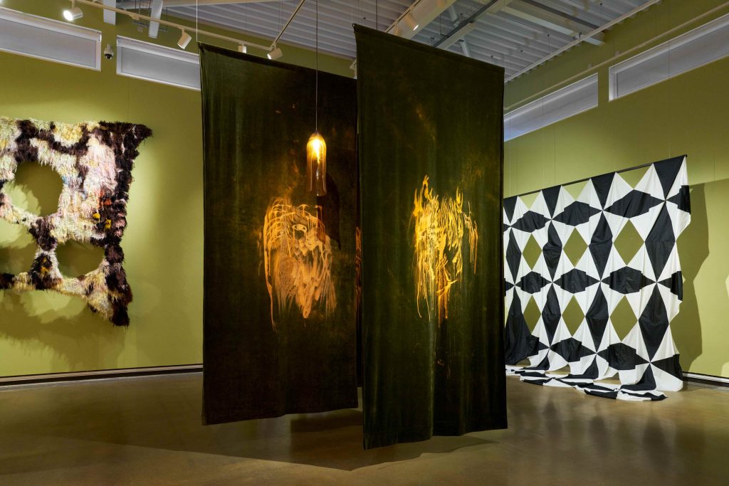 To the left is a giant tufted wall hanging of brown and white yarns. In the middle are large swaths of brown velvet hanging from the ceiling with burnt out natural circles in the middle, with a lamp in between the two fabrics. To the right is a large-scale black and white piece made of attaching many umbrella fabrics together, that pool onto the floor.
