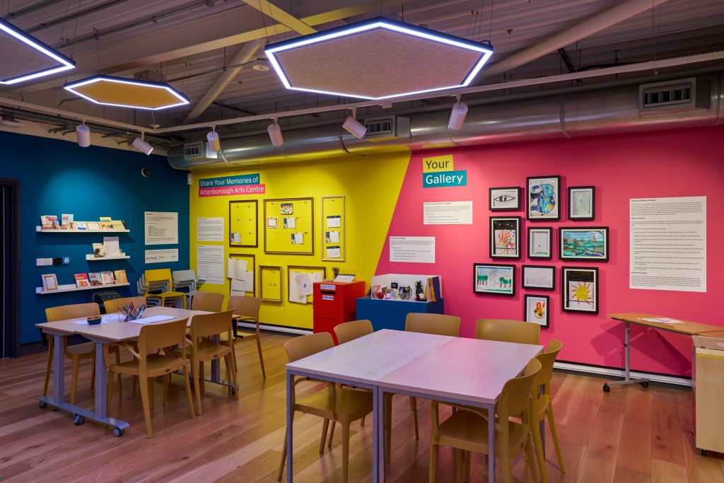 An art room with blue, yellow and pink walls covered with interactive elements on the walls to showcase artwork. In the room are tables and chairs filled with art materials.