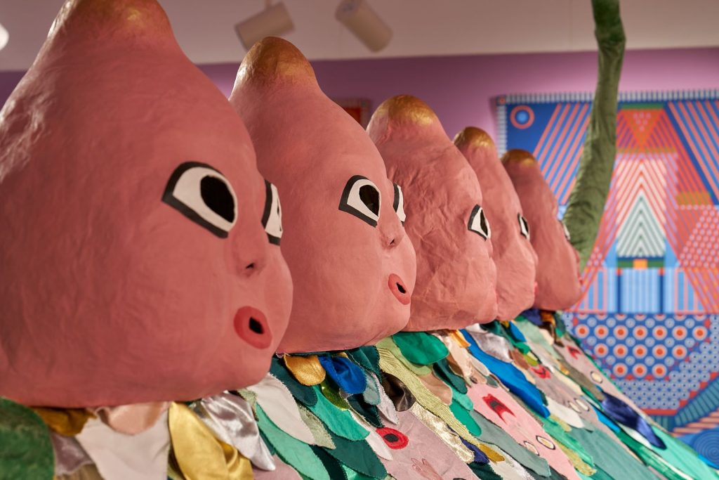 Five pink, paper mache, cartoon heads in a line looking outward.