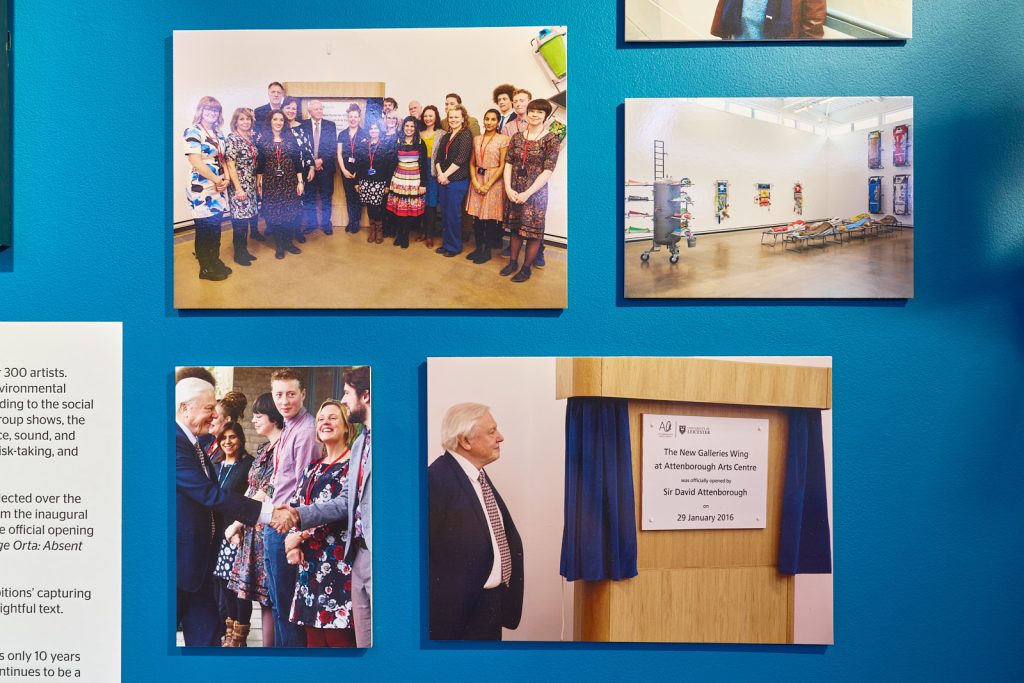 A blue wall with four photos showing the Attenborough Arts Centre team 10 years ago stood in the gallery as it opened, the exhibition itself, the outside with David Attenborough greeting crowds as he walks in, and David Attenborough stood next to a plaque commemorating the opening of the gallery.