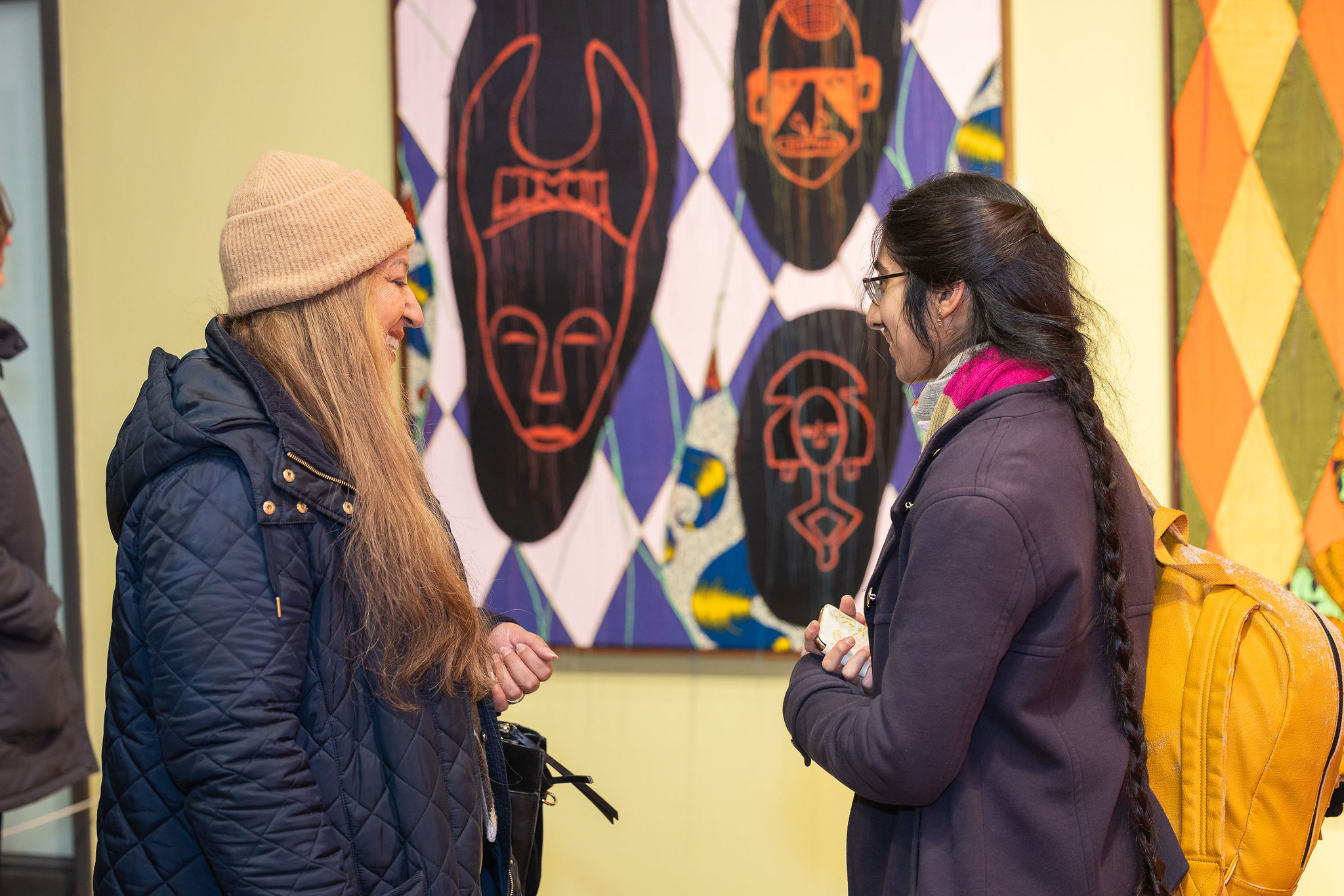 Two women in a gallery in front of art made of fabric on canvas, chatting happily together.