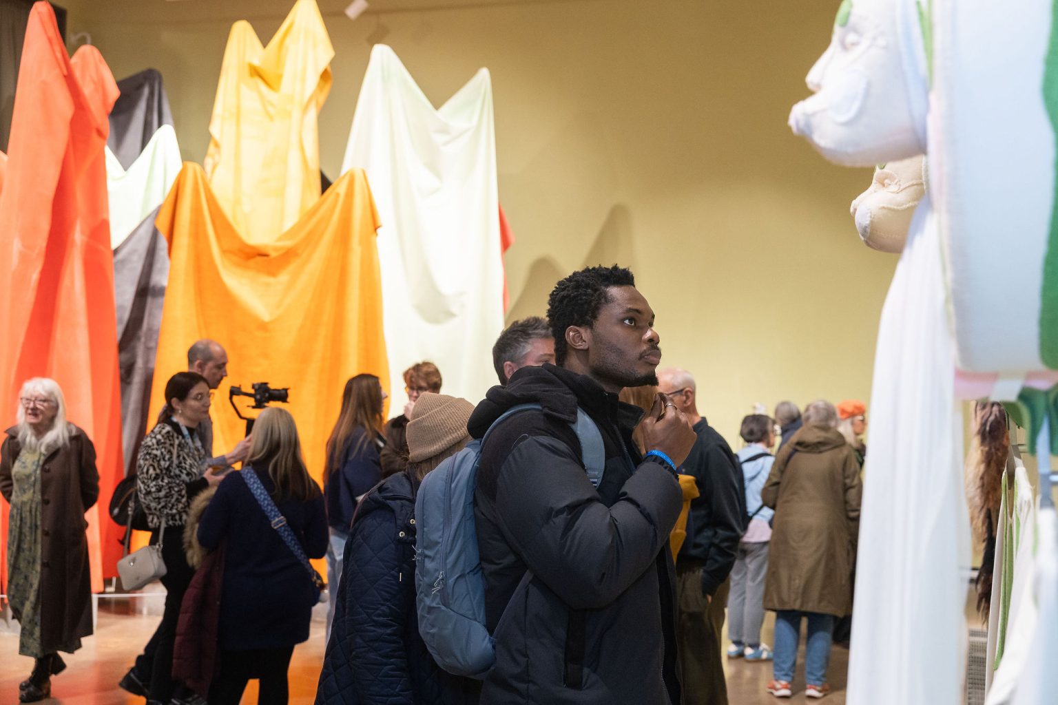 A room full of people looking at sculptures of large piece of fabric hanging from poles.