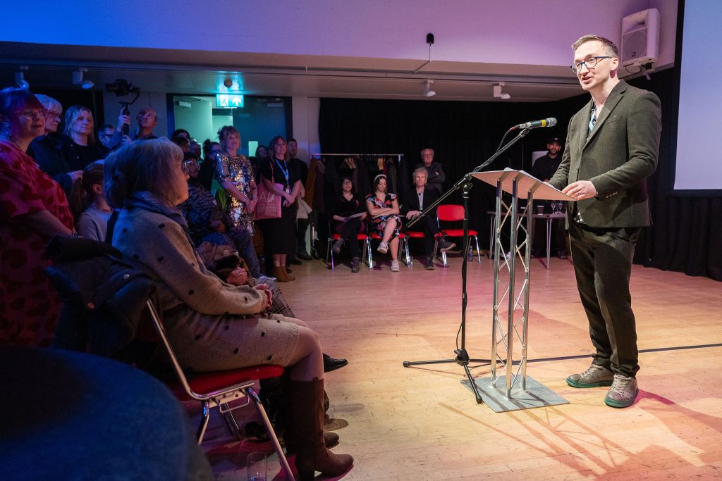 An audiences to the left watching a man to the right speak into a microphone.