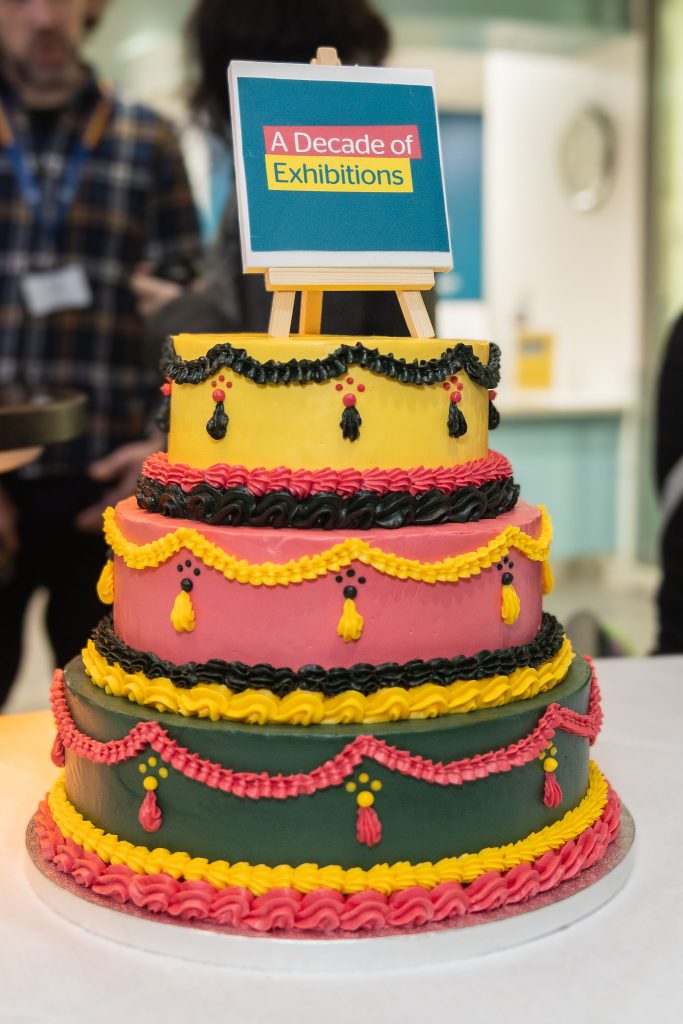 A multi coloured, three tiered cake with an easel on top saying 'A Decade of Exhibitions'.
