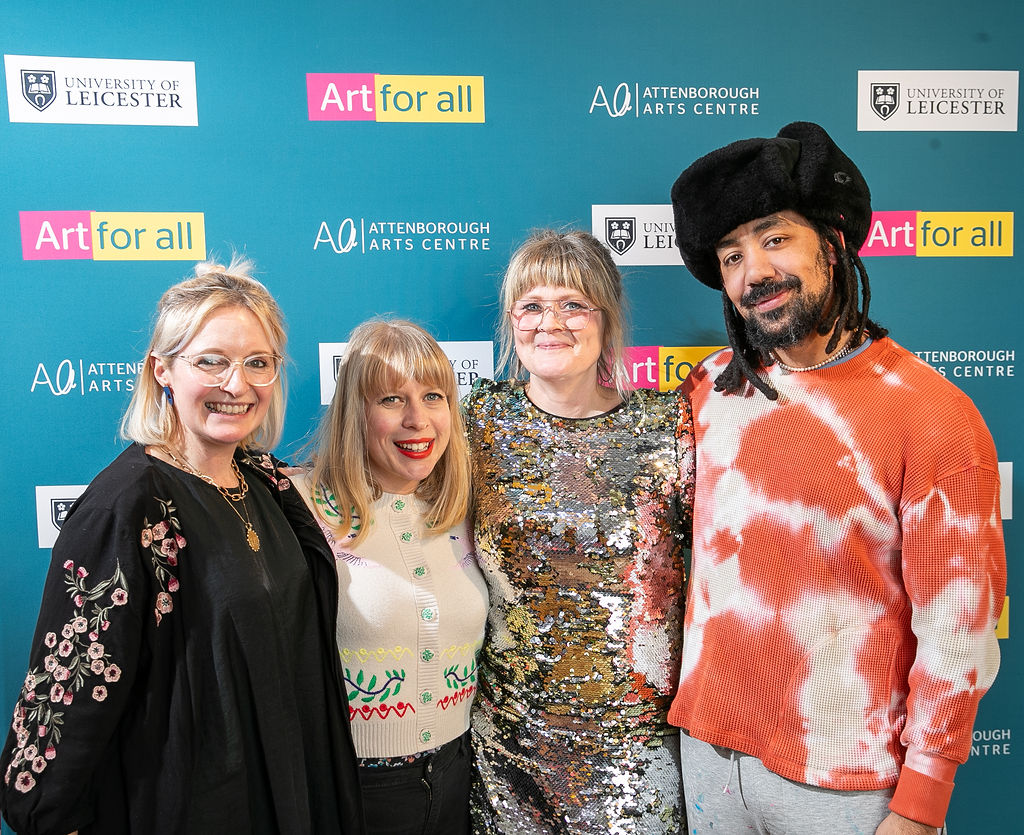Three white women and a black man wearing party clothing and smiling together in front of a board with logos.