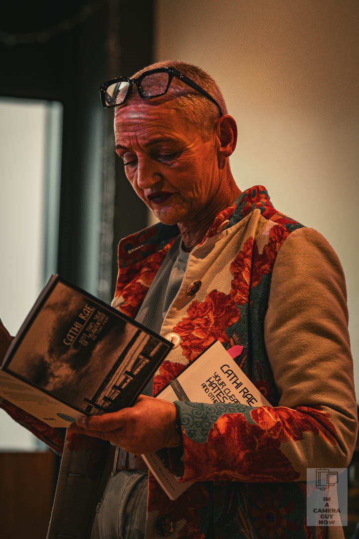 An older white woman with a shaved head reading a book in a darkened room.