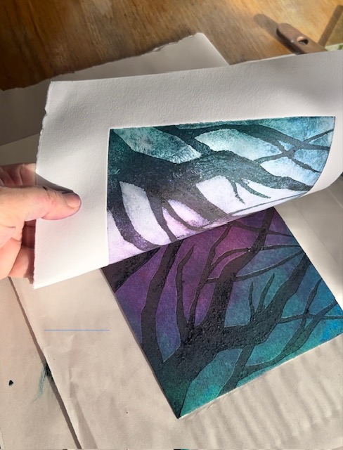 A piece of paper being lifted off of a printed block to reveal a purple, blue and black print of a tree.