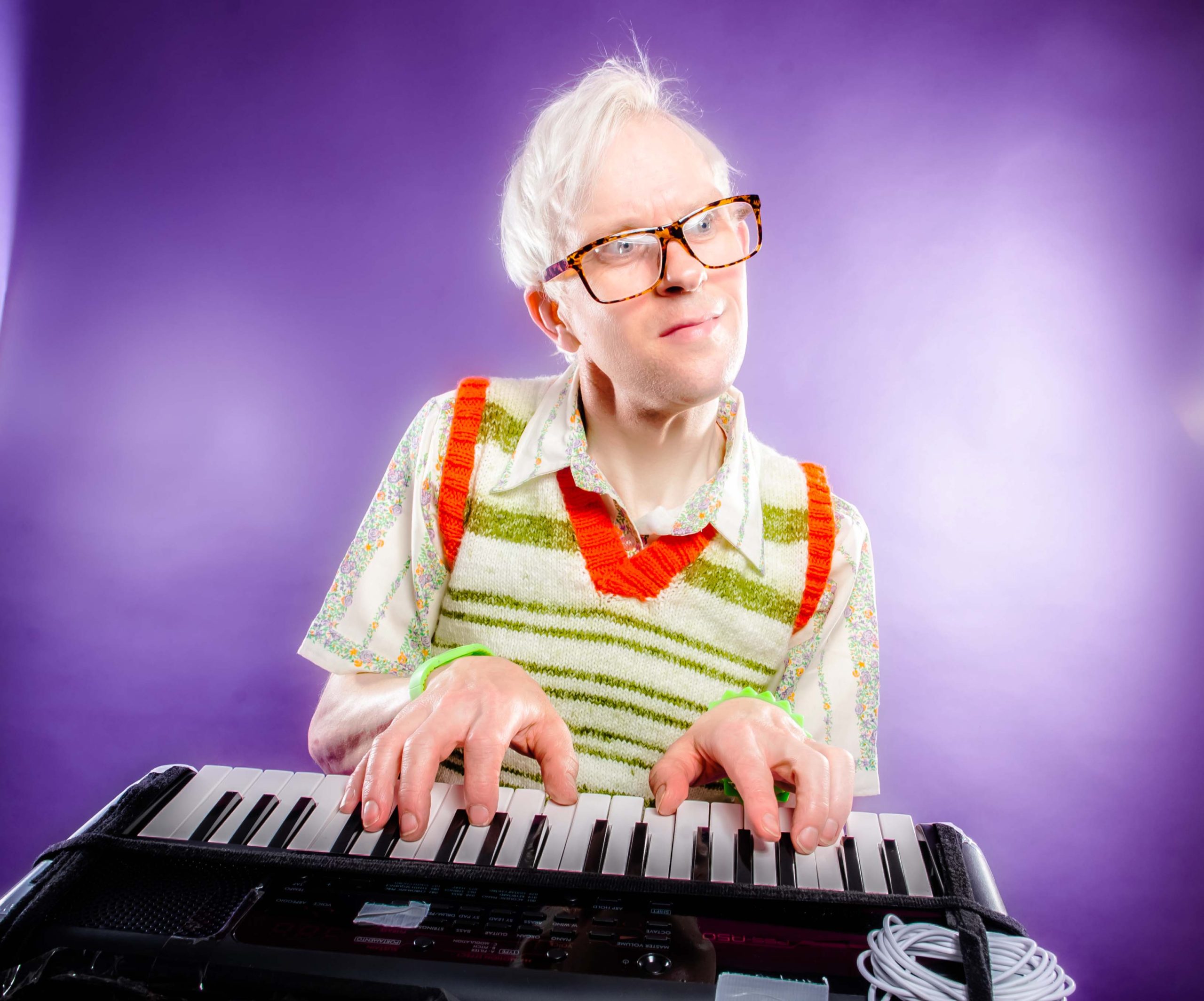 A white haired white man wearing glasses and a sweatervest, playing the keyboard in front of a purple background.
