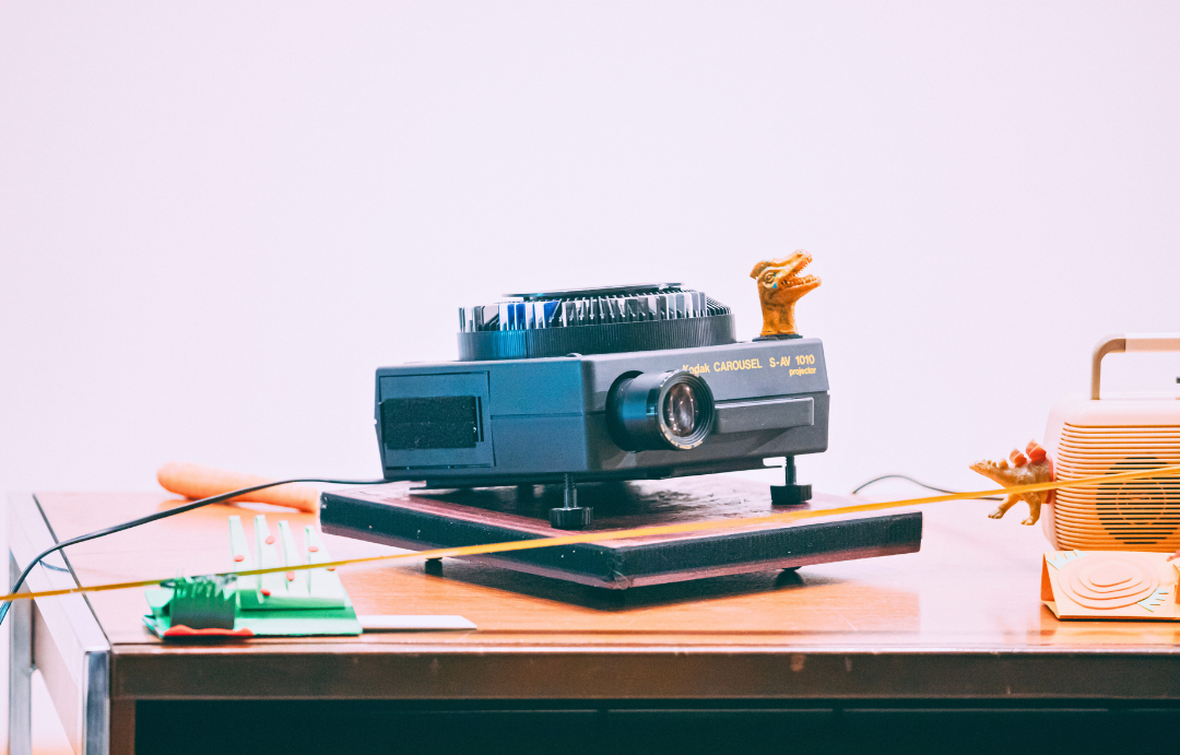 An old projector on a table with a toy dinosaur head on top of it positioned in the corner