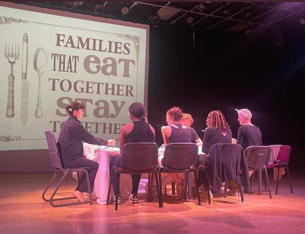 A group of people, possibly actors, are in a hall sat around a table with a projection on the screen in front of them that says "families that eat together stay together."