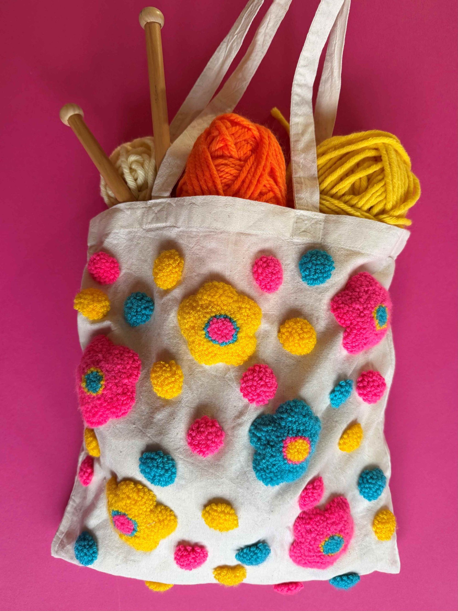 A tote bag embellished with punch needle embroidery. Balls of yarn and punch needles are overfilled and flowing out of the bag.