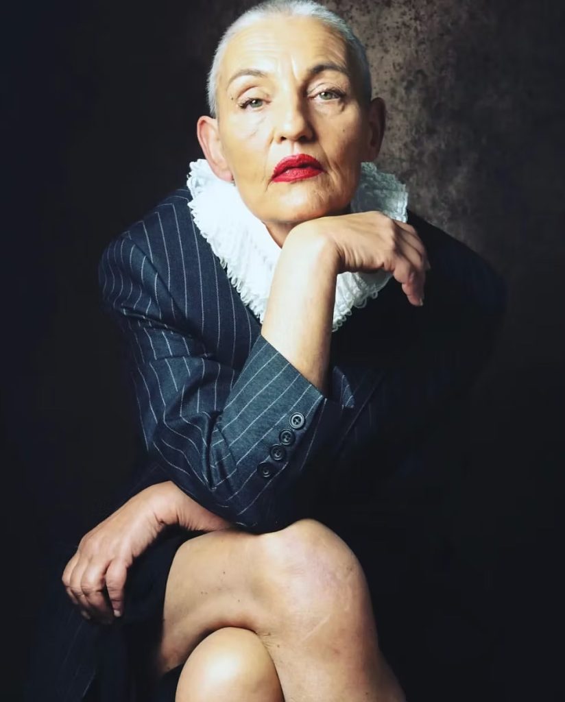 An older white woman with a shaved head and red lipstick, resting her chin on her hand while sitting crossed legged in a blazer and neck ruff.