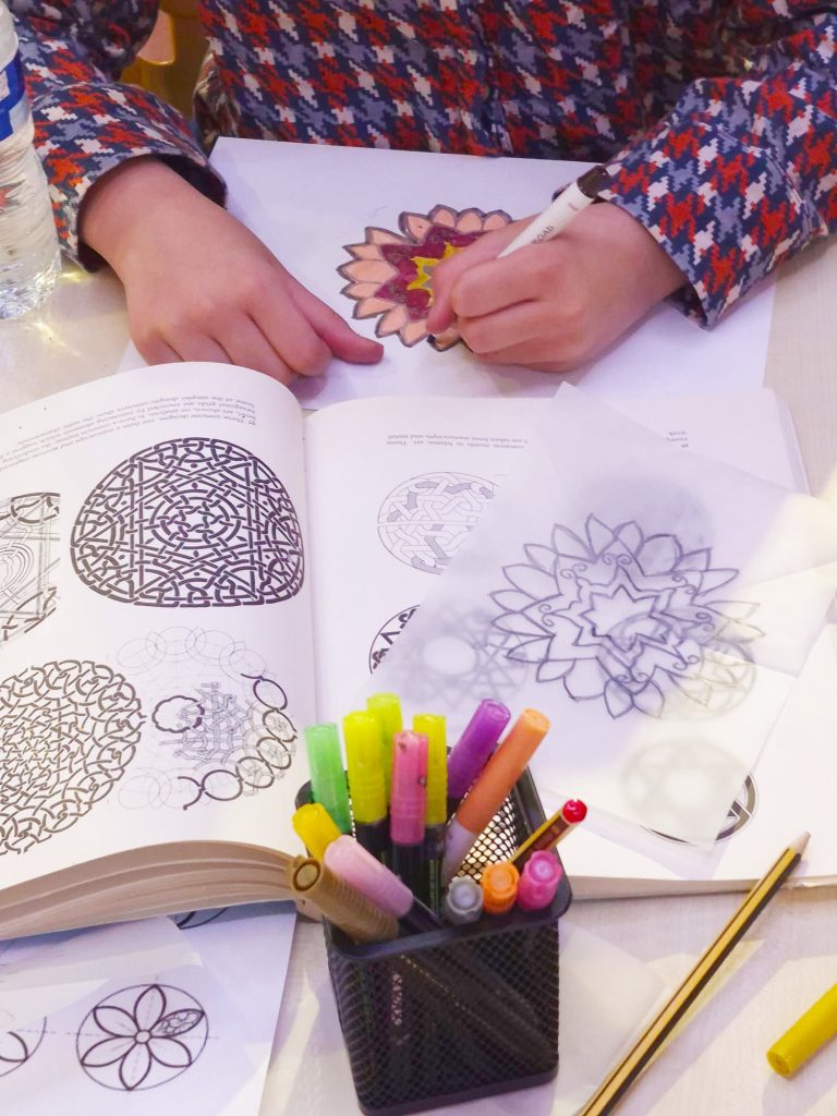 Two hands drawing out Islamic Geometry symbols with books in front of them showing different patterns.