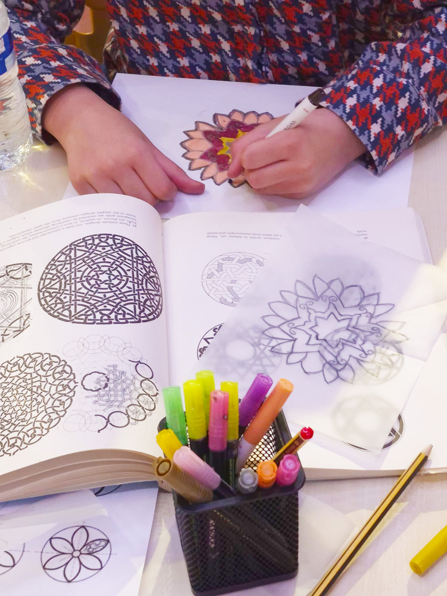 Two hands drawing out Islamic Geometry symbols with books in front of them showing different patterns.