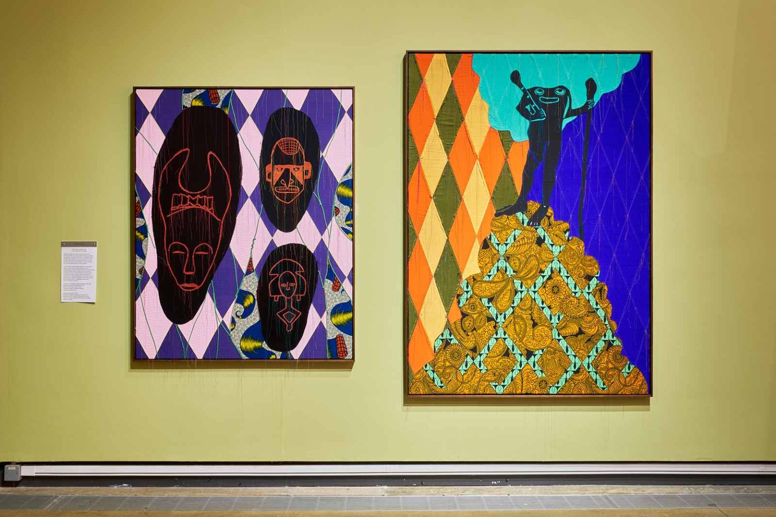 Two canvas artworks on a wall. The left is made of fabric in a purple and pink harlequin pattern with black shapes and red outlines of African style masks. The right piece has various patterns showing the sky and a hill, with a frog-like man stood atop the hill looking at a cloud with a staff in hand.