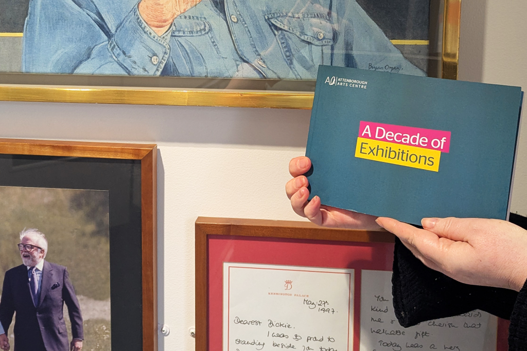 A hand holding a book with the title 'A Decade of Exhibitions' in front of framed pictures of Lord Richard Attenborough.