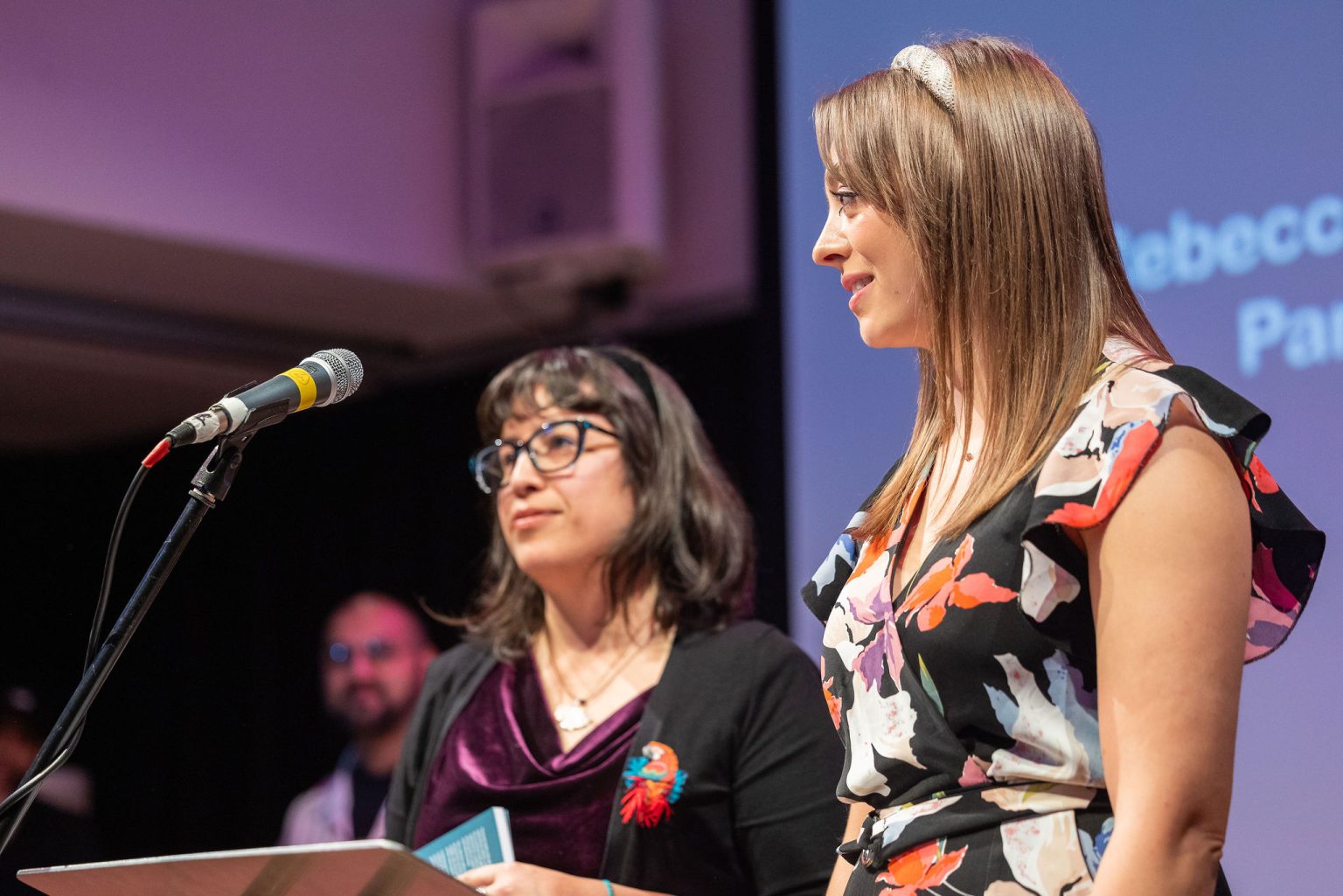 Two woman stood in front of a microphone speaking to an audience.