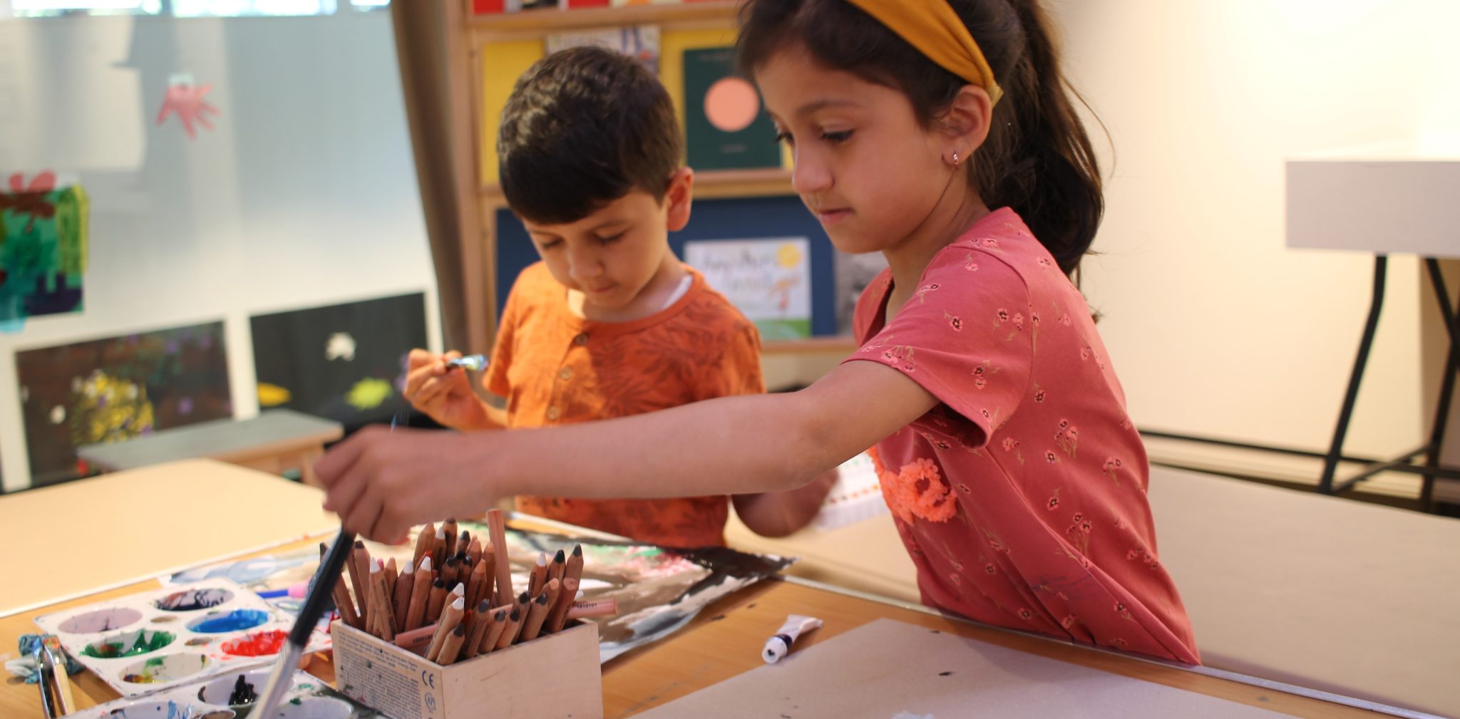 Two young children are at a table with paints, drawing onto paper.