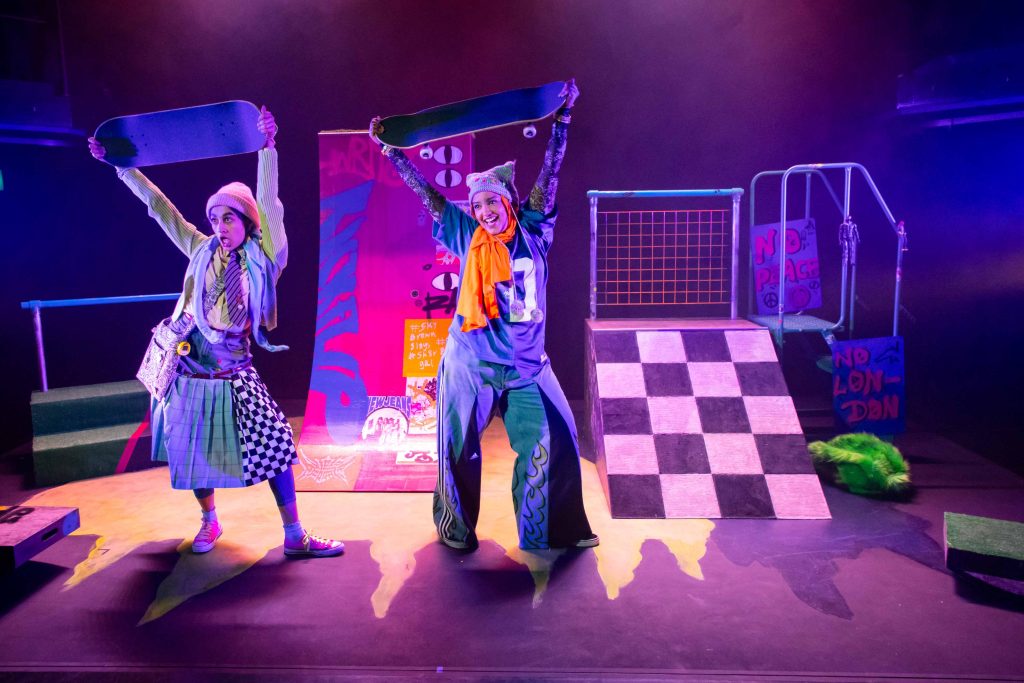 Two brightly dressed women are on a stage holding up skateboards above their heads. The background set it bright and colourful with skatepark ramps.