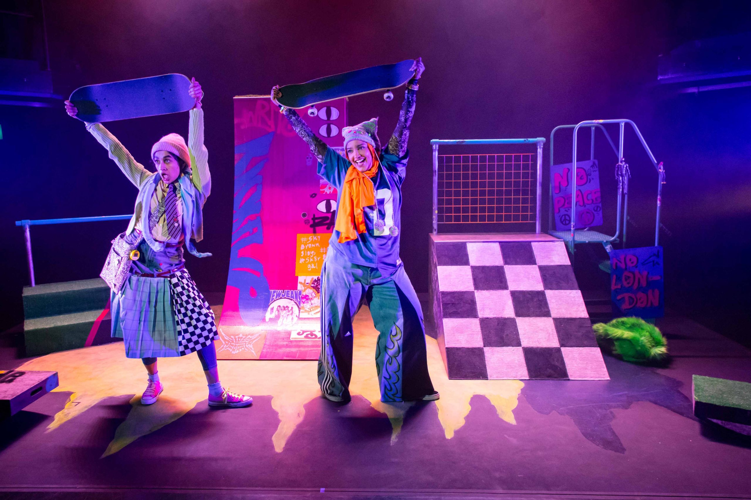 Two brightly dressed women are on a stage holding up skateboards above their heads. The background set it bright and colourful with skatepark ramps.