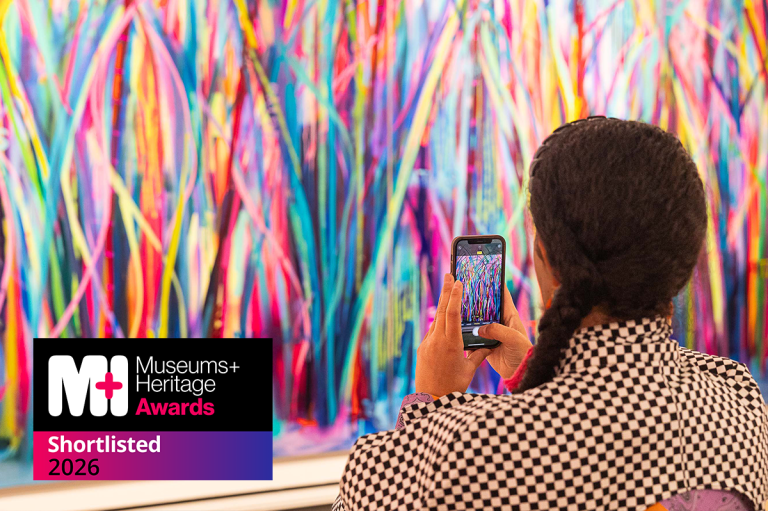 The back of a woman holding up her phone to take a picture of the artwork in front of her. The artwork is bright colourful lines in large-scale.