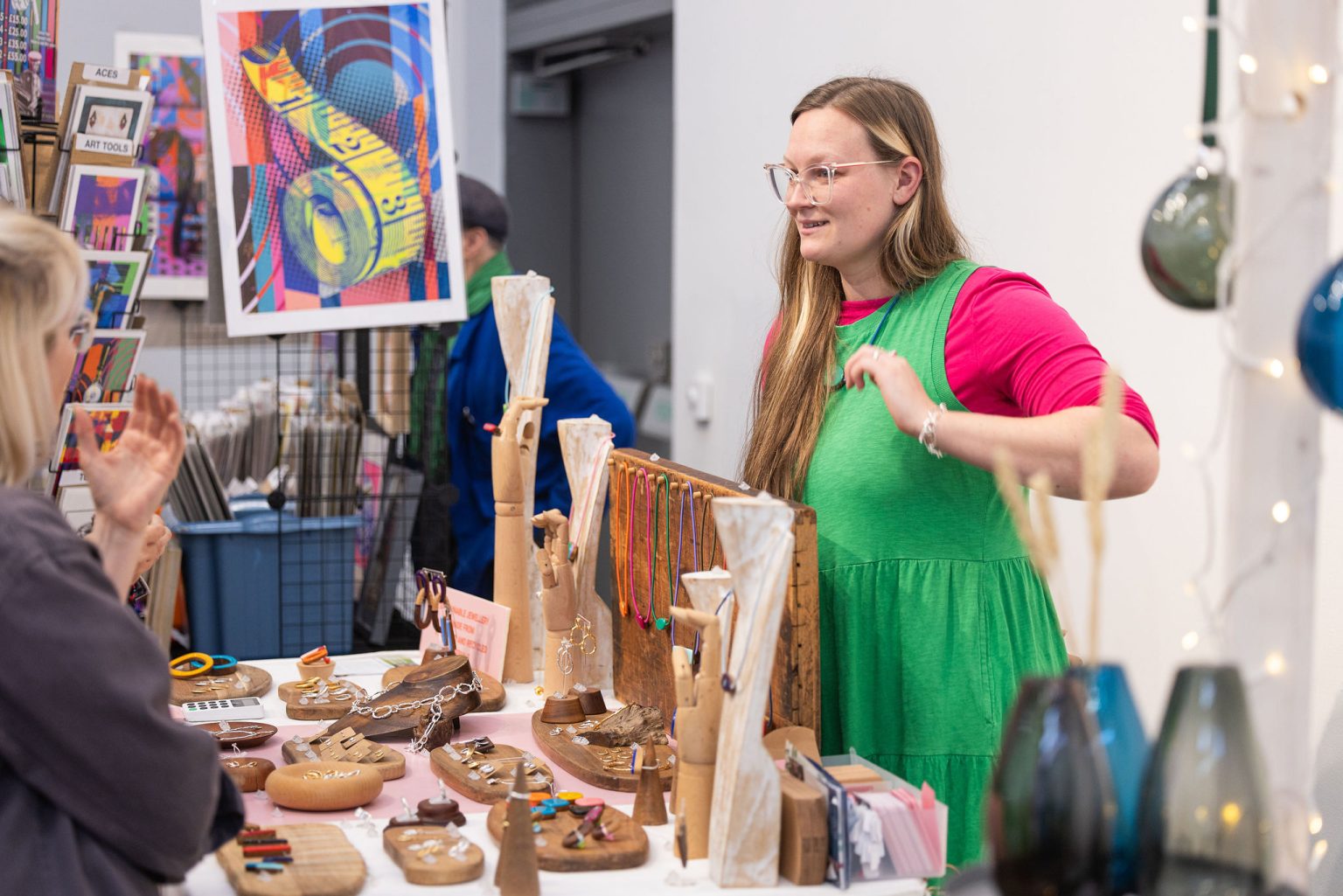 A white woman is bright colourful clothing stood behind a stall with jewellery displayed talking to a customer.