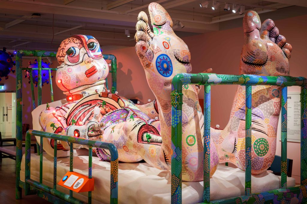 A large sculpture of a catoonish character covered in drawings lying on a hospital bed.