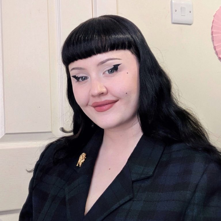 A white woman with long black hair, a short fringe and heavy eyeliner smiling at the camera.
