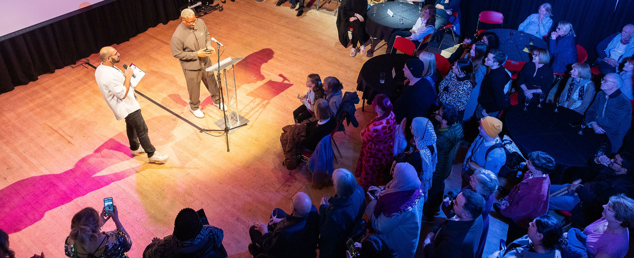 Two people stood in a spotlight to the left speaking into a microphone to an audience on the right bathed in blue.