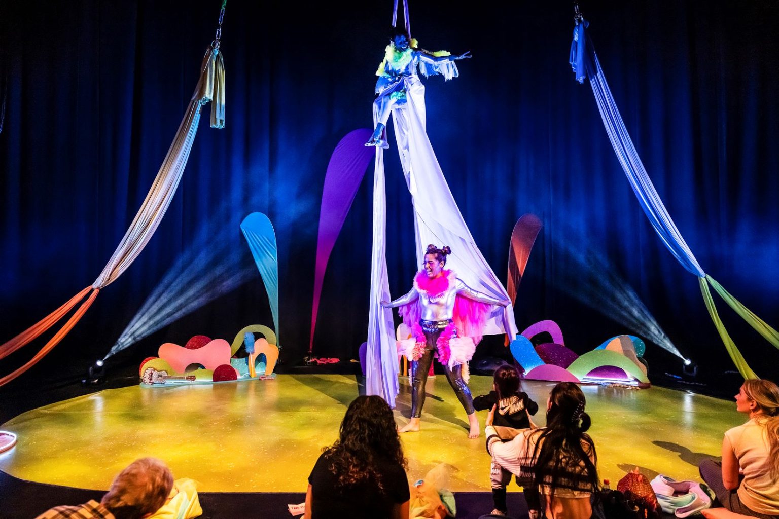 A colourful stage with an audience watching two women in theatrical clothing on aerial ropes.