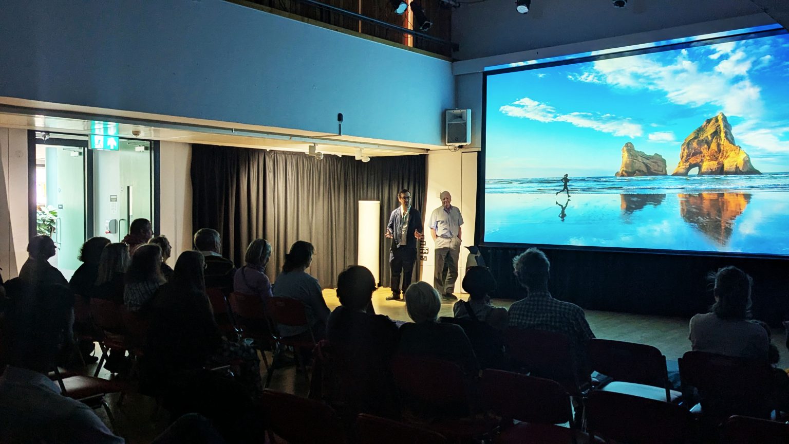 An audience sat down in a dark room, looking at a large screen showing a blue beach. To the left a man speaks next to a cardboard cut-out of David Attenborough.