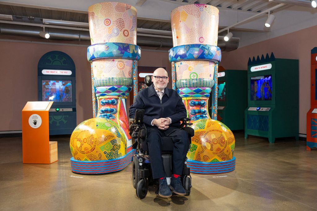 A white middle-aged man in a wheelchair smiling in front of a pair of giant colourful boots and legs with braces, covered in swirling patterns.