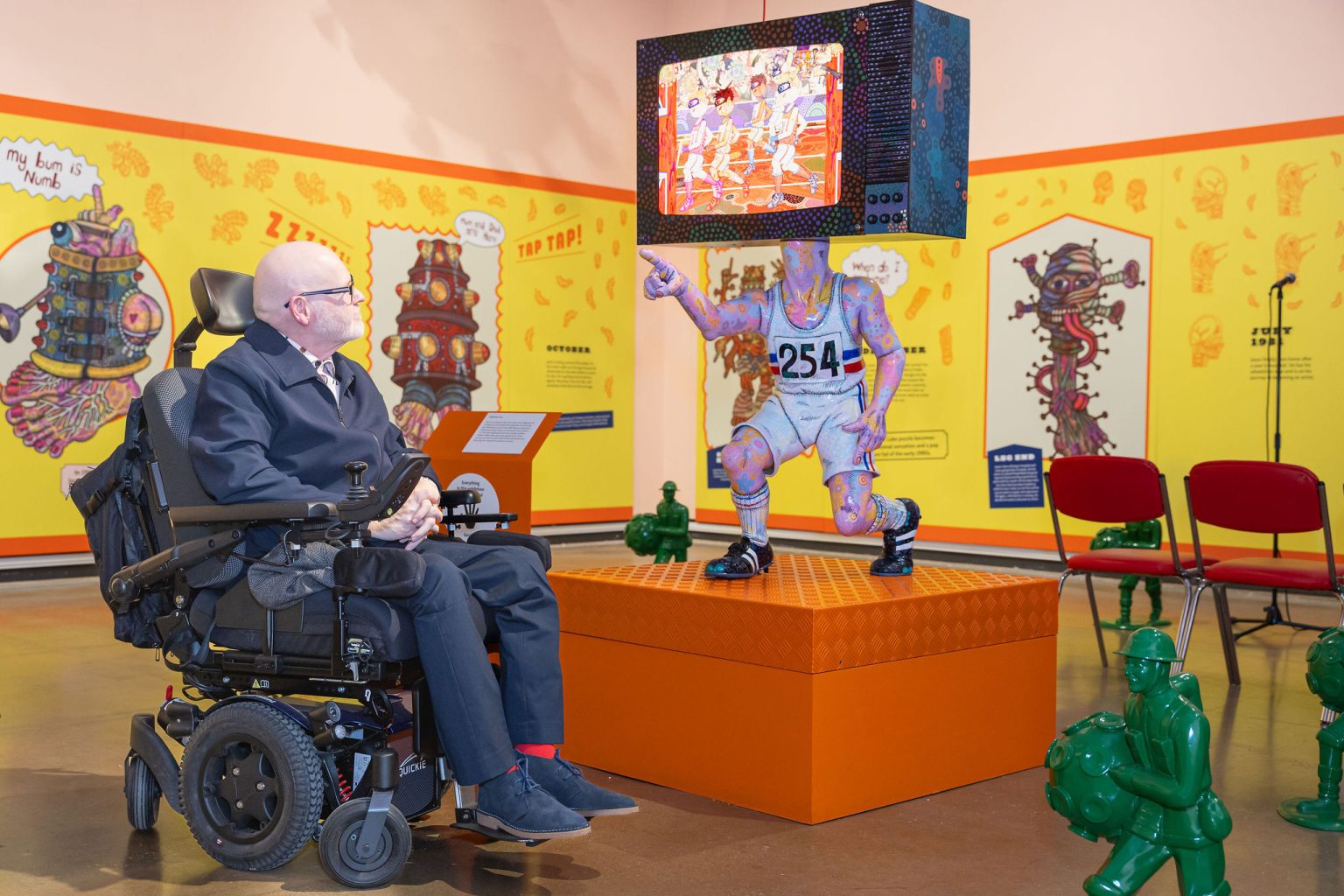 A white middle-aged man in a wheelchair in a colourful gallery space, looking up at a colourful sculpture of a man in running gear with a tv head pointing forward.