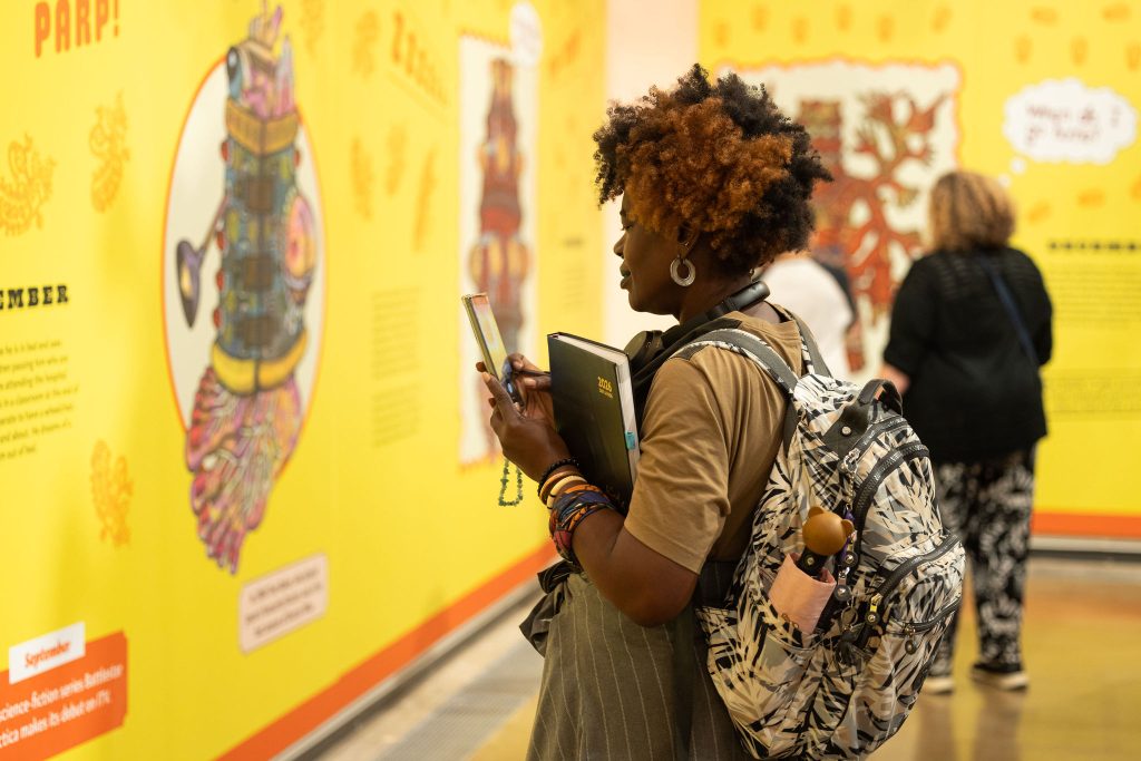 A black women with an afro taking a picture of a colourful yellow wall with giant illustrations.