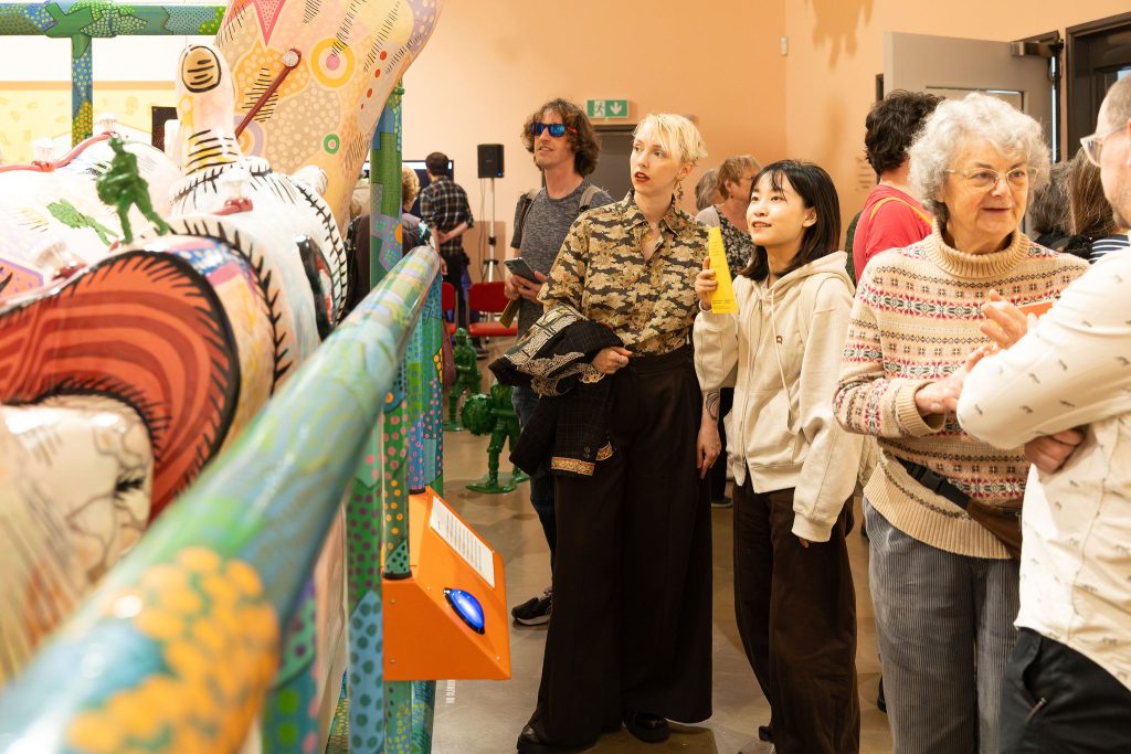 A group of people looking to the left at a giant colourful sculpture of a person in a hospital bed, covered in colourful swirls.