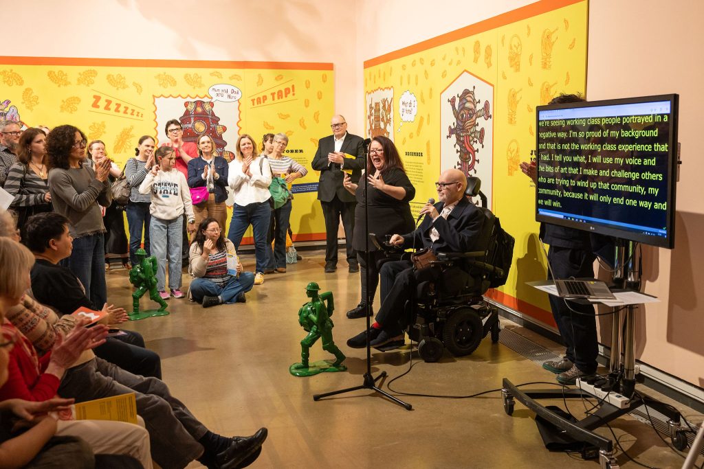 Jason Wilsher-Mills; in front of a crowd in a gallery give a speech with a microphone. To the right is a tv screen showing the closed captioning text.